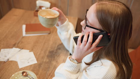 lady wearing glasses and white sweater answers phone call while holding ceramic latte cup in cozy cafe, seated at wooden table with napkin sketches and empty saucer in soft ambient light