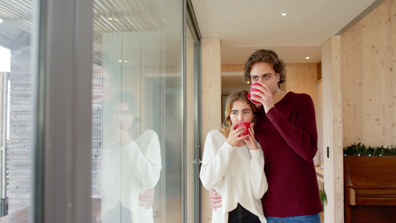 Christmas time, Couple enjoying cozy moment at home, holding red mugs by window, copy space