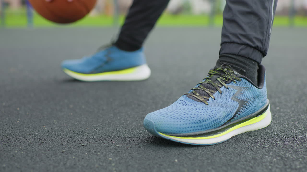 Scarpa da ginnastica blu su campo in asfalto, giocatore di streetball che si allena con il palleggio, primo piano sulla tomaia in maglia, suola neon, lacci testurizzati, angolazione dal basso, pallone da basket che rimbalza, sfondo di parco urbano grintoso