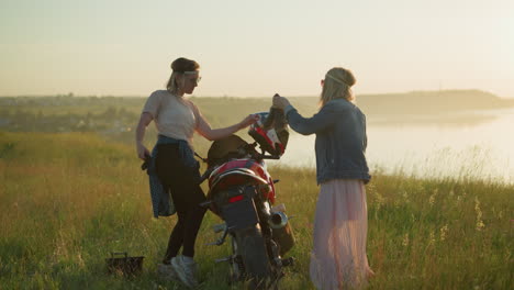 two friends happily dancing with cleaning rags near a red motorcycle in an open grassy field with a serene river in the background