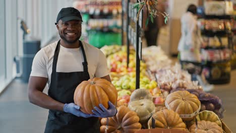 Retrato-De-Un-Hombre-Feliz-De-Piel-Negra-Con-Una-Camiseta-Blanca-Cerca-De-Una-Vitrina-Con-Calabazas.-Un-Hombre-Sostiene-Una-Calabaza-En-Sus-Manos,-Posa-Y-Sonríe-Ampliamente-En-Una-Gran-Tienda-De-Comestibles.