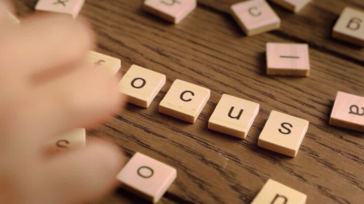 A hand arranges wooden scrabble-style tiles to spell the word focus on a rustic wooden surface.