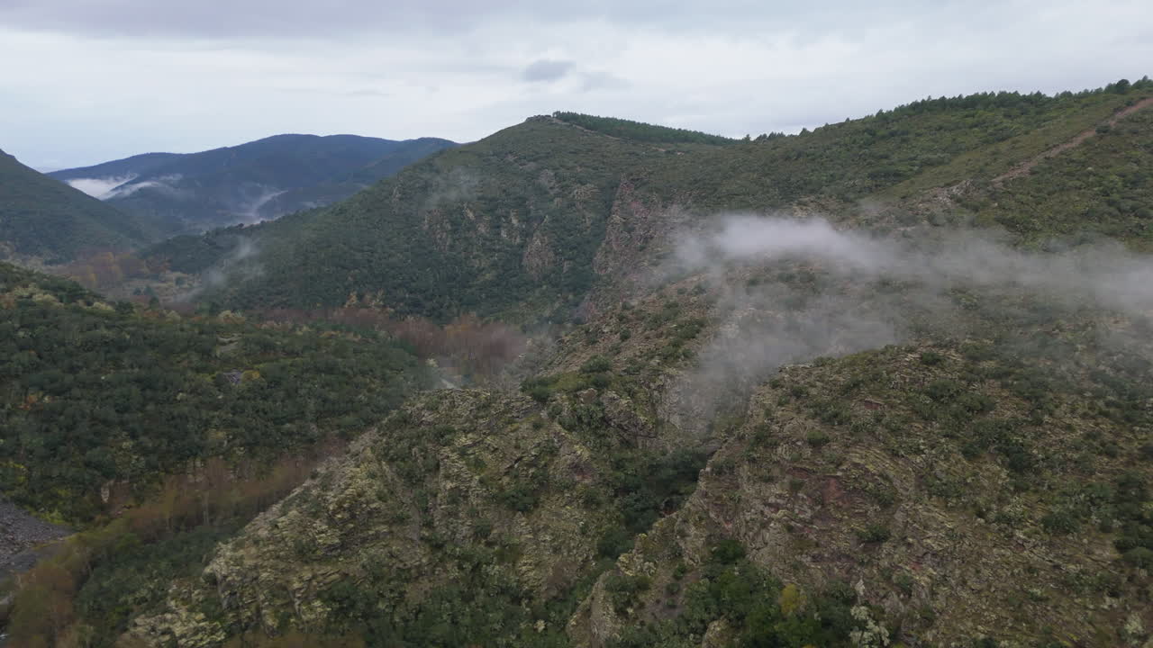 Drone footage of rugged mountain terrain on a cloudy day, with low mist and clouds floating across the green hillsides, creating a mysterious and cinematic mood