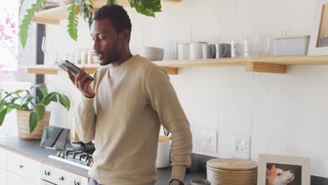 happy african american man leaning on countertop in kitchen, talking on smartphone