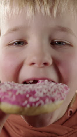 boy with satisfied expression bites into glazed pink donut, frontal vertical