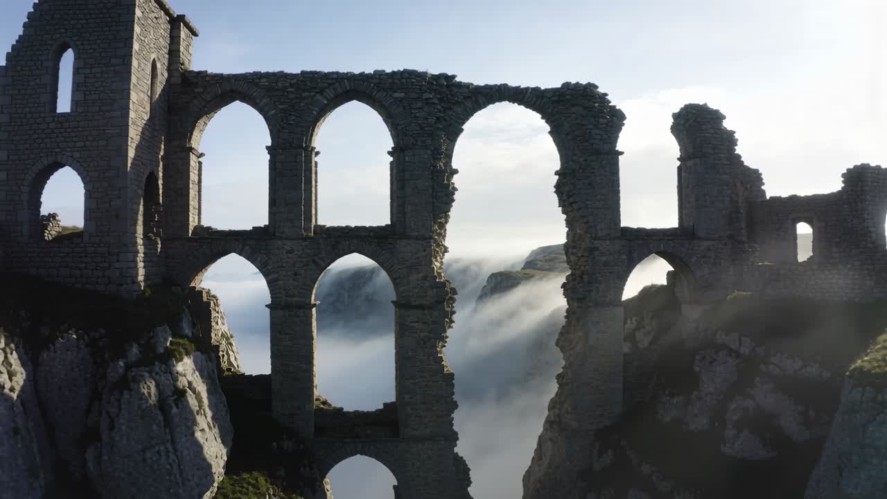 La telecamera di un drone si allontana, rivelando una rovina su una gola con una torre, archi crollati e nebbia che si sposta.