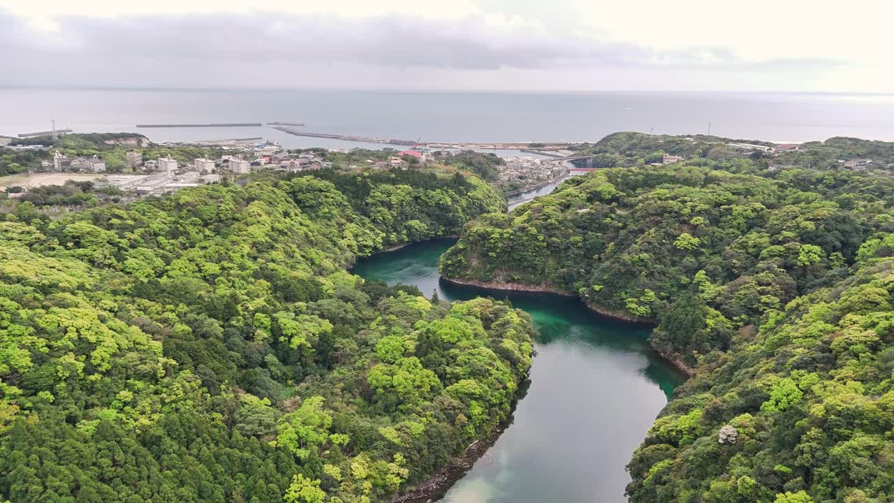 Aerial view over Matsumine Bridge, Yakushima, Japan with dense green forest, winding river, and the coastal harbor extending toward the open sea under cloudy skies, blending nature with settlement