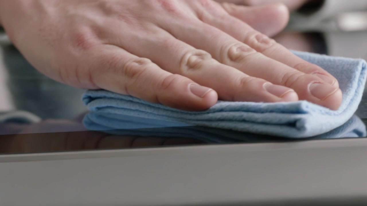 Eine wischende Hand in grauem Ärmel nähert sich dem Kochfeld mit einem gefalteten hellblauen Tuch und entfernt Flecken.