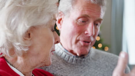 Cerca-De-Una-Pareja-Mayor-Sentada-En-Un-Sofá-En-Casa-Charlando-Con-Un-árbol-De-Navidad-En-Segundo-Plano.