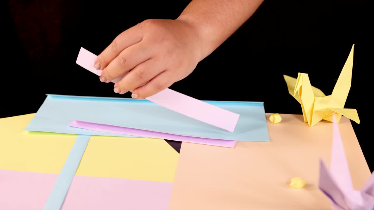 Hands cut pastel paper strips with scissors on colorful table; origami cranes, studio lighting visible