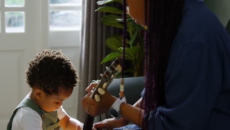 Vista-Lateral-De-Una-Joven-Madre-Negra-Y-Un-Hijo-Pequeño-Tocando-La-Guitarra-En-El-Salón-De-Una-Cómoda-Casa-4k