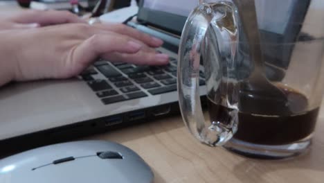 detailed close-up shot of a glass coffee mug with a spoon, and hands actively typing on a laptop