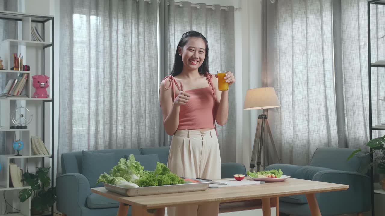 미소 짓는 아시아 여성 이 오렌지 주스 한 잔 을 들고 집 에서 건강 한 음식 을 준비 하는 동안 엄지손가락 을 올려 놓은 몸짓 을 하고 있다