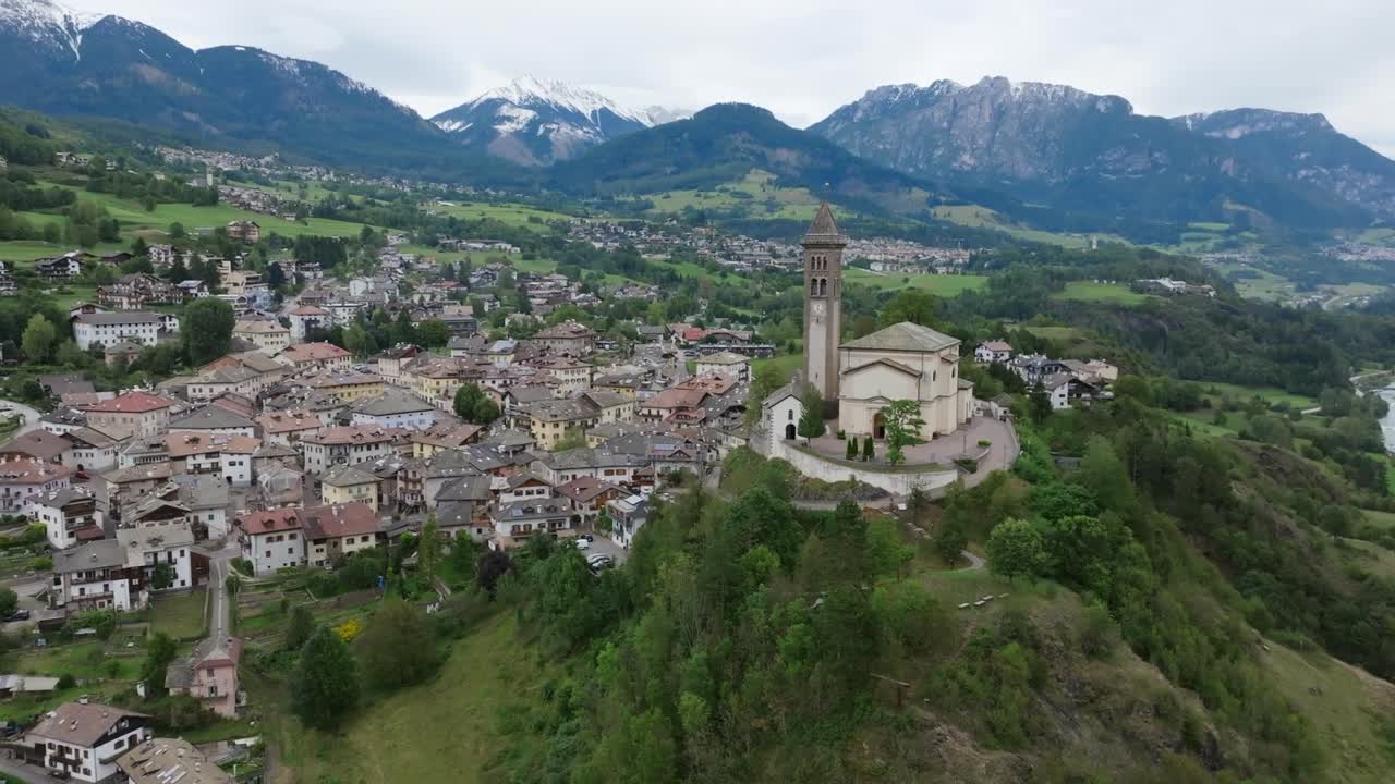 Drone view of historic hilltop church with circular stone wall above alpine village in Trentino-Alto Adige surrounded by forest and mountains