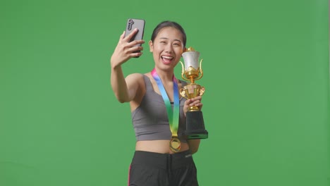 asian woman with a gold medal holding a gold trophy and taking photo on smartphone as the first winner on green screen background in the studio