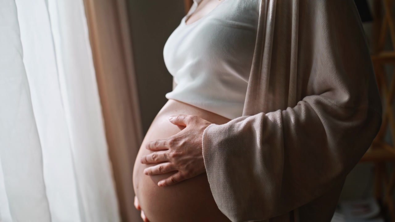Donna incinta che accarezza il pancione vicino alla finestra. Primo piano di una madre che tocca il pancione