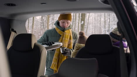 camera focuses from inside a car to a man offers a hot drink from a thermos to her wife and daughter while they talking