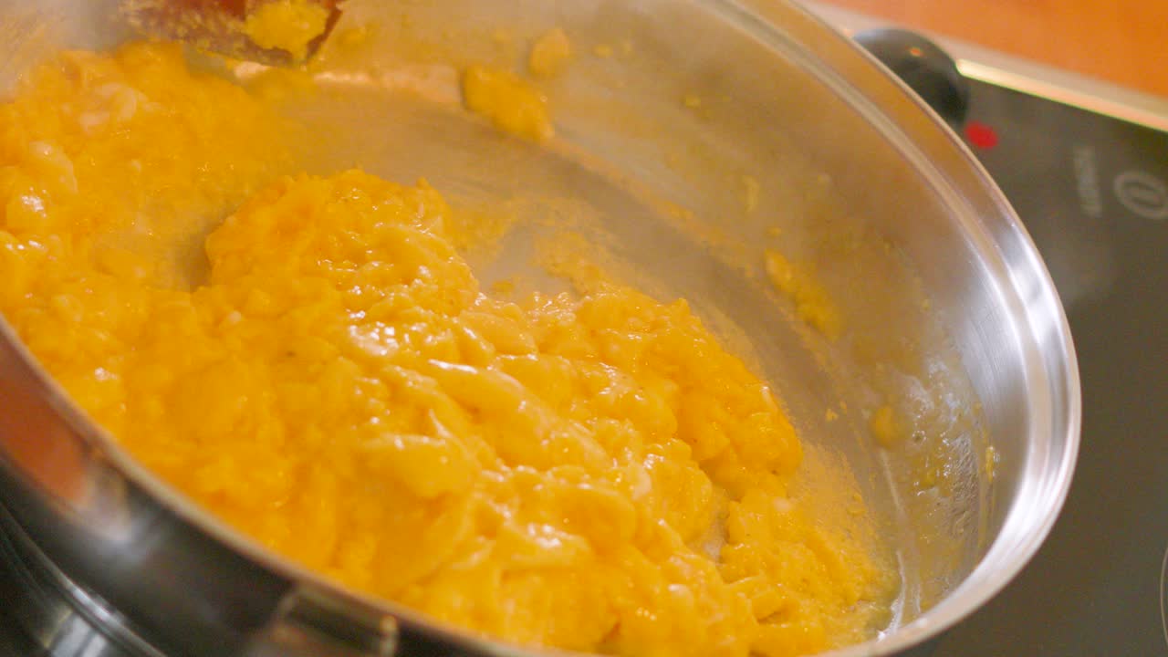 Skewed Side Angle of Golden Scrambled Eggs Being Gently Cooked in Pan by Chef with Wooden Spatula with Hot Steam Rising in Slow Motion. Cooking Hot Food Footage.