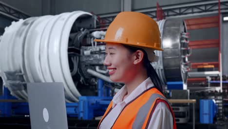 close up side view of asian female engineer with safety helmet working on a laptop and looking around while standing with airplane engine maintenance conducted
