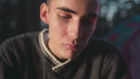 close up young man wearing dark sweater eating canned food with knife, seated outdoors near glowing tent, turns and smiles while looking at someone in background