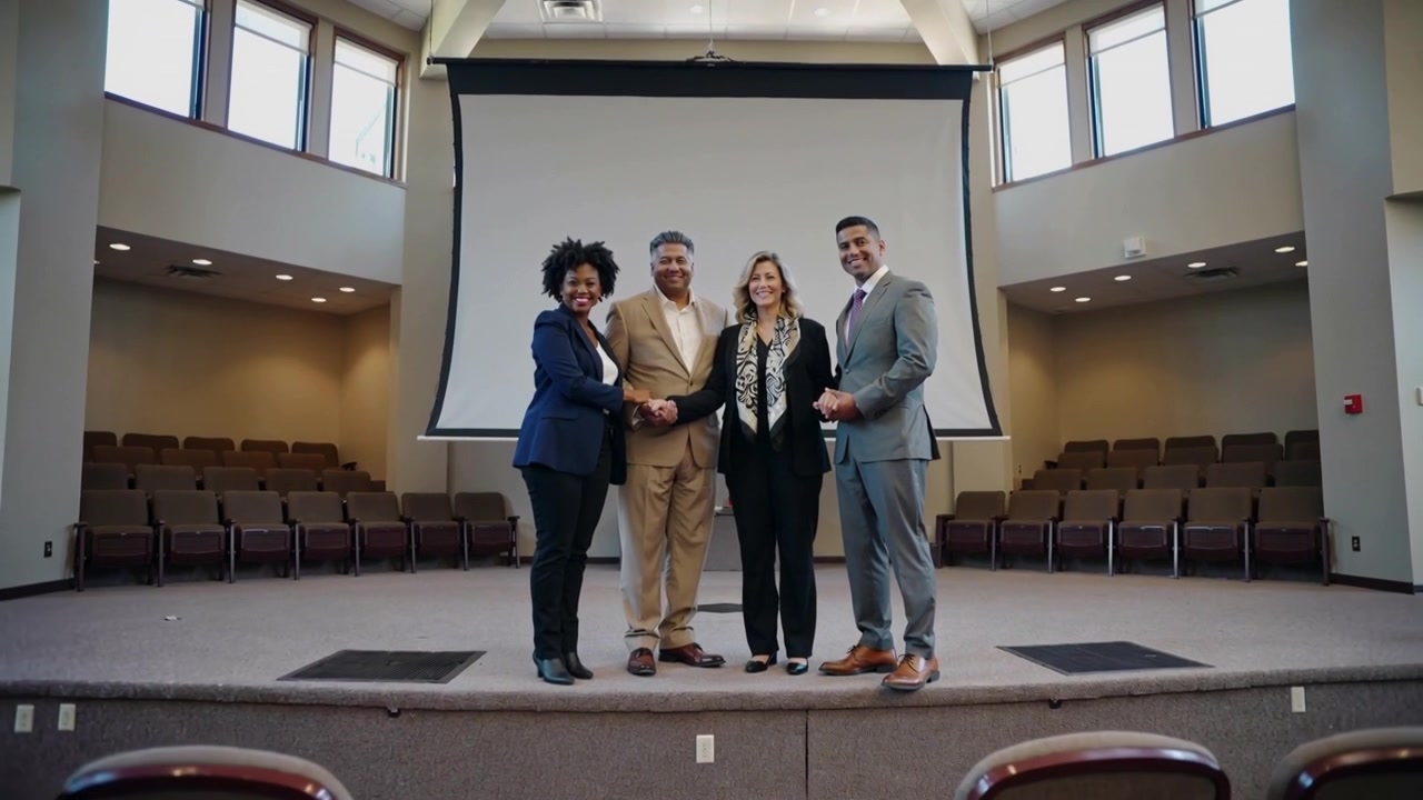 Responding to cue, four presenters in suits stepping on stage, linking hands near projection screen
