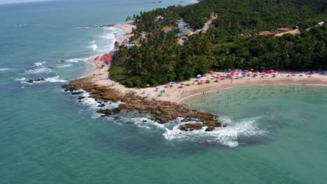 Tilt-down-rotating-aerial-drone-shot-of-the-popular-tropical-Coquerinhos-beach-surrounded-by-palm-trees-and-covered-in-umbrellas-with-tourists-swimming-in-a-natural-pool-in-Conde,-Paraiba,-Brazil