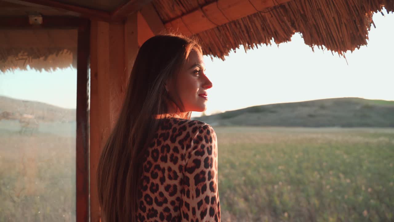 Caucasian brunette long hair pretty woman wearing stylish animal pattern dress in wooden lookout tower looks at rural landscape in countryside, handheld close up portrait