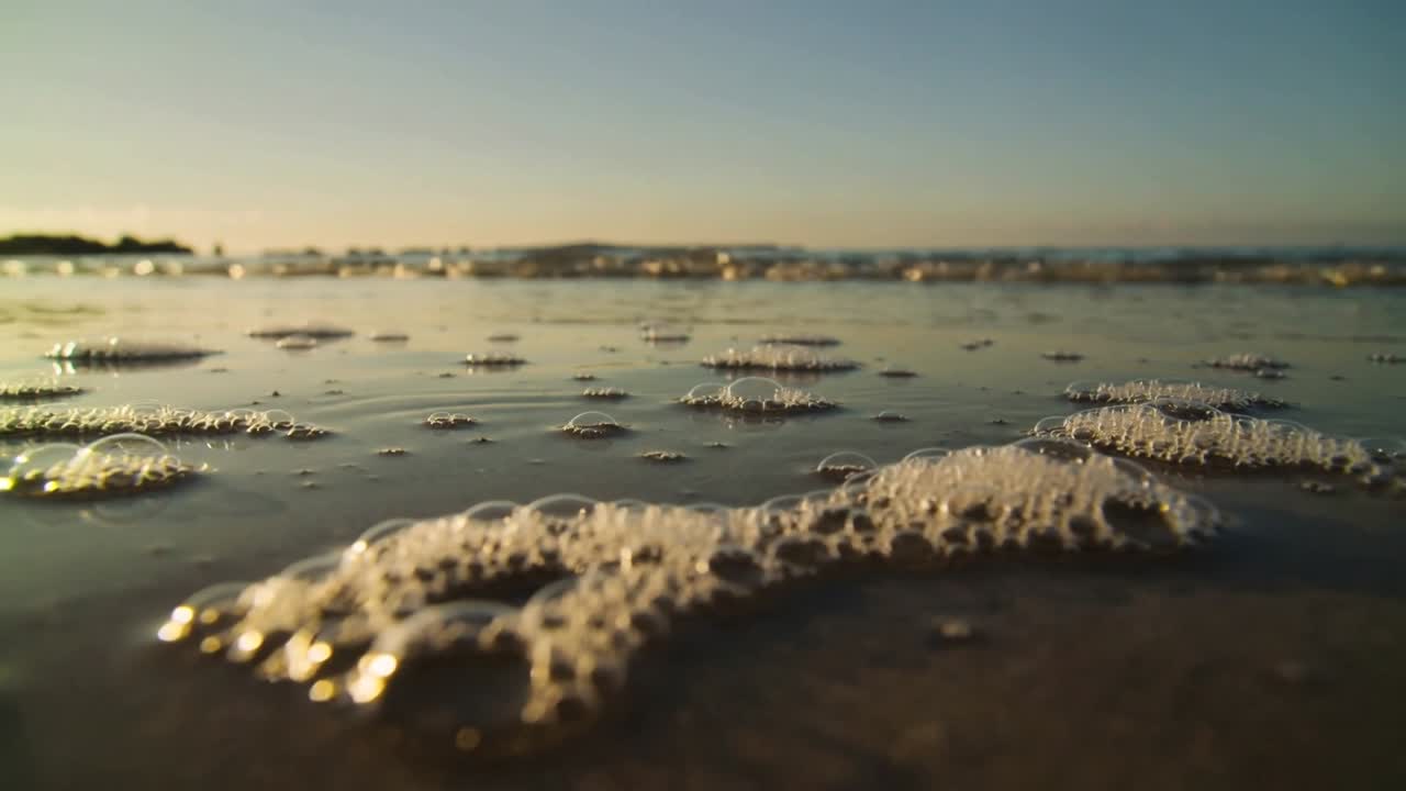 Scena naturalistica con ammassi di schiuma marina che si spostano sulla sabbia bagnata, con un'onda in arrivo che forma bolle.
