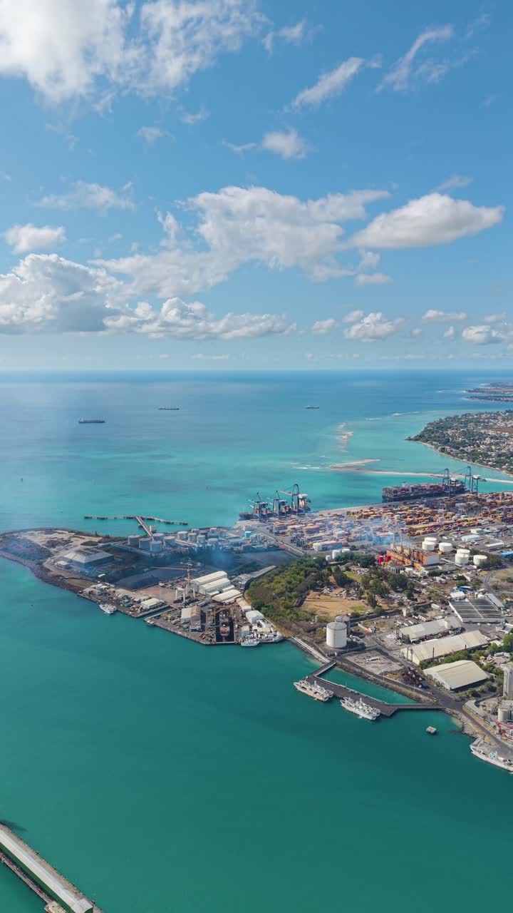 Vertical aerial of the Port Louis industrial harbor and container terminal in Mauritius. The shot captures a busy commercial port with cranes, cargo ships, and containers by the turquoise ocean