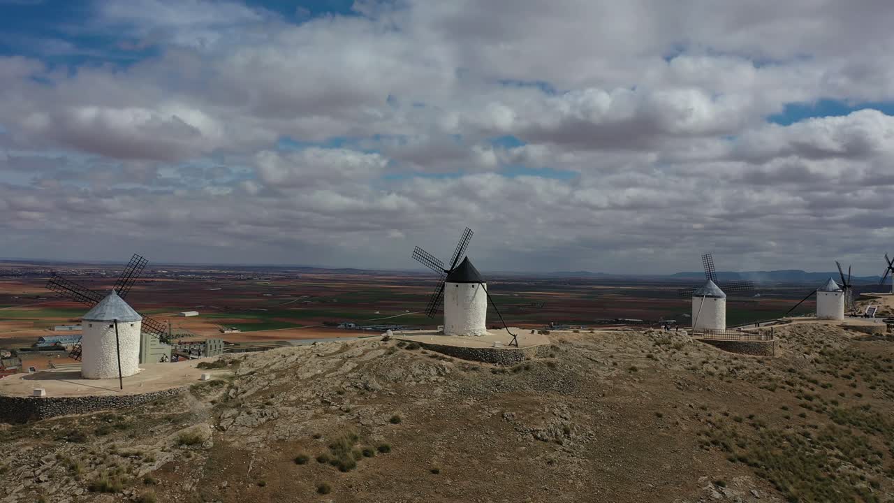 vuelo lateral con dron visualizando cuatro viejos molinos de viento visitados por personas que hacen turismo con un fondo de tierras de cultivo y población, en un día nublado de primavera en toledo, españa