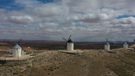 lateral flight with drone visualizing four old windmills visited by people doing tourism with a background of farmland and population, on a cloudy spring day in toledo-spain