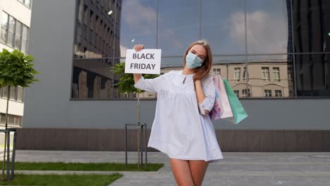 girl in protective mask with shopping bags showing black friday inscription during coronavirus