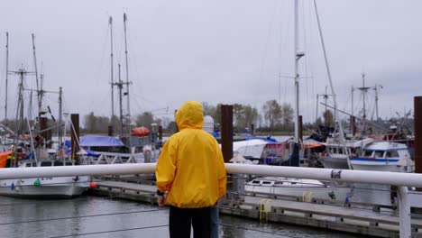 Medium-Shot-of-a-person-in-a-yellow-jacket-walks-dockside-overlooking-a-pier-of-fishing-boats