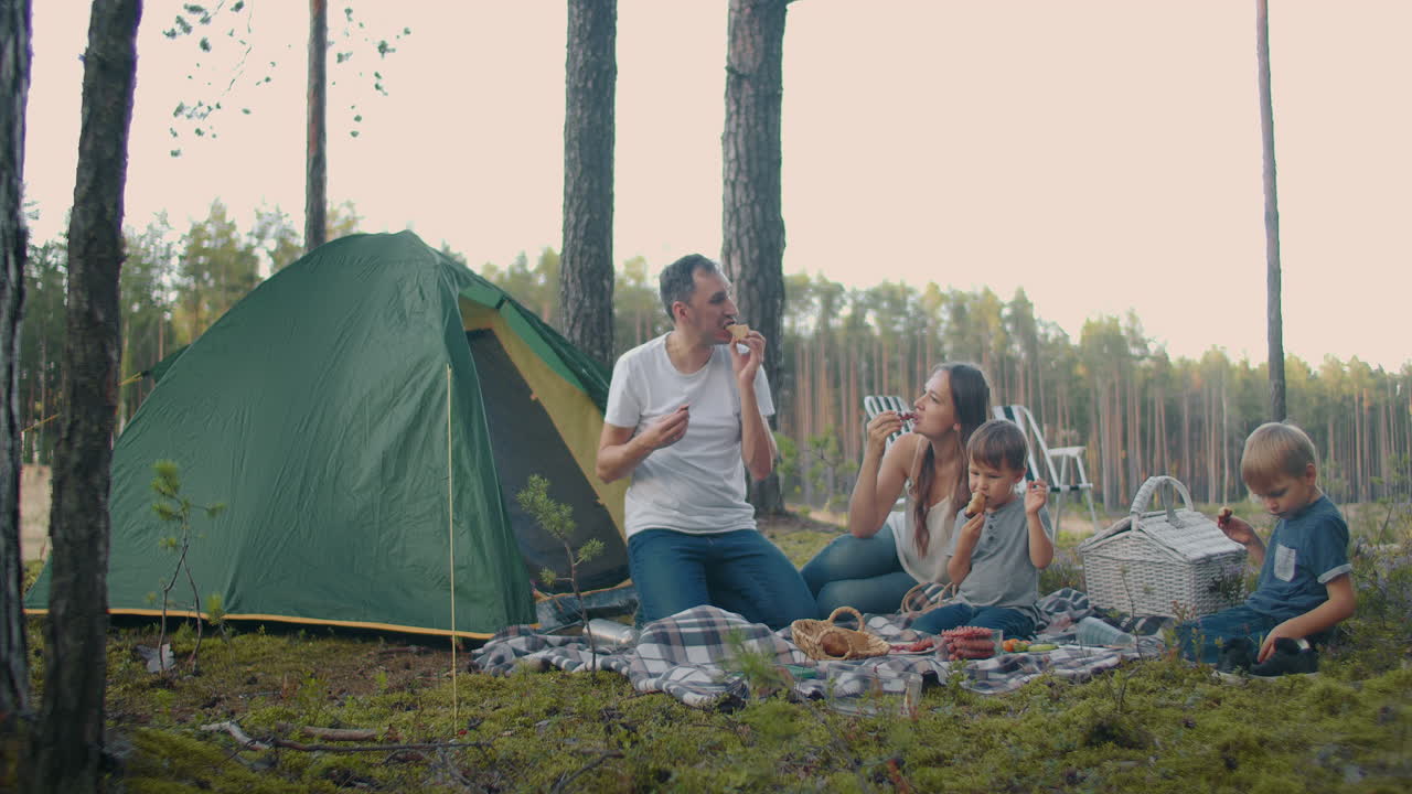 자연에서 아이들과 함께 가족의 피크닉 부모와 두 작은 아들은 숲 캠프에서 풀에 앉아 먹고 있습니다.
