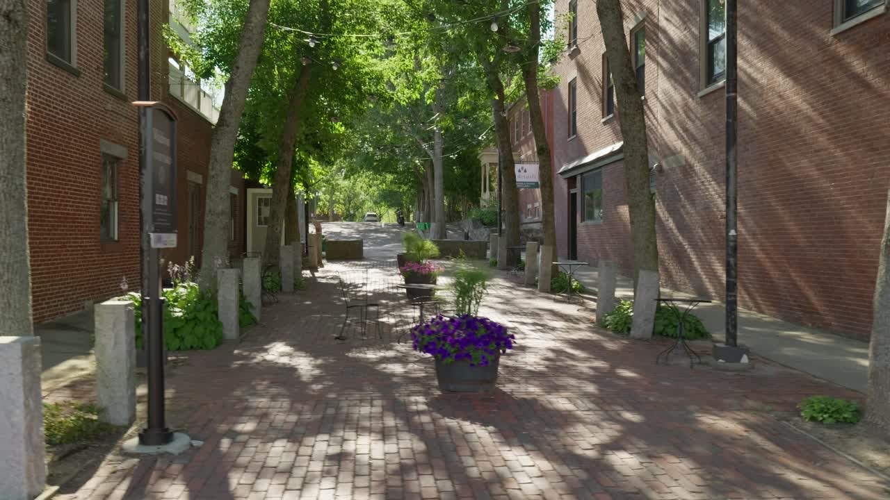 En langsom gåtur ved en lille murstensterrasse med potteblomster og træer, hvor eftermiddagssolen filtrerer igennem. Den flankeres af to røde murstensbygninger.