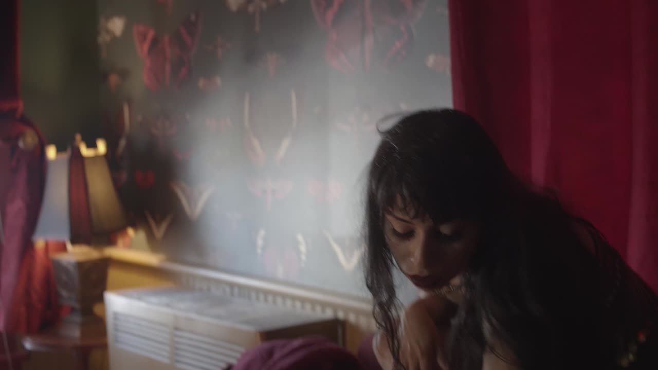 una mujer joven con un aspecto gótico visualmente y lentamente se inclina hacia adelante con una expresión seductora en una habitación retro oscura con una cortina roja