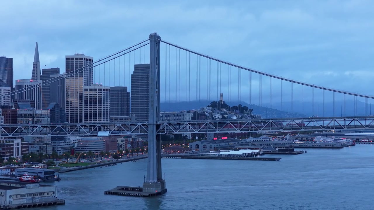 Video drone che orbita attorno al San Francisco Bay Bridge all'ora blu, rivelando lo skyline illuminato, l'acqua calma e un cielo crepuscolare suggestivo. Ampio paesaggio cinematografico cattura la bellezza del paesaggio urbano