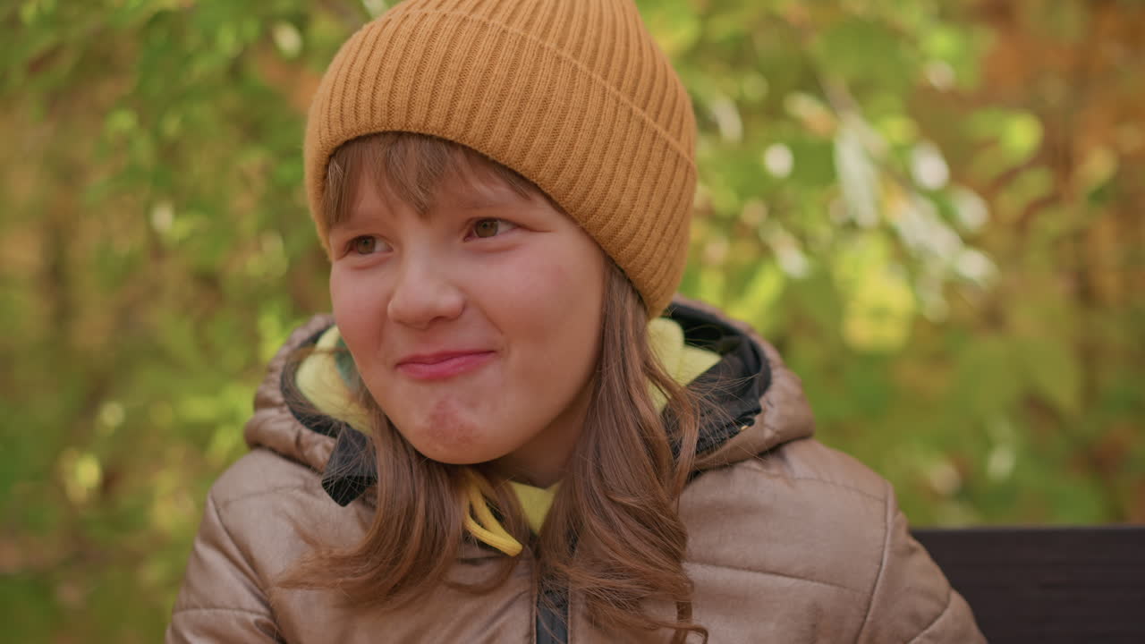 close up of cheerful child in brown beanie and padded jacket sitting on bench smiling while eating, surrounded by blurred autumn foliage, warmth and innocence of childhood
