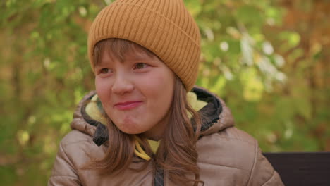 close up of cheerful child in brown beanie and padded jacket sitting on bench smiling while eating, surrounded by blurred autumn foliage, warmth and innocence of childhood