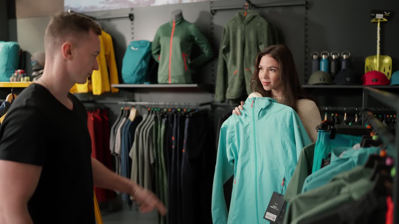 The girl in the store asks the guy's advice choosing between two sport jackets