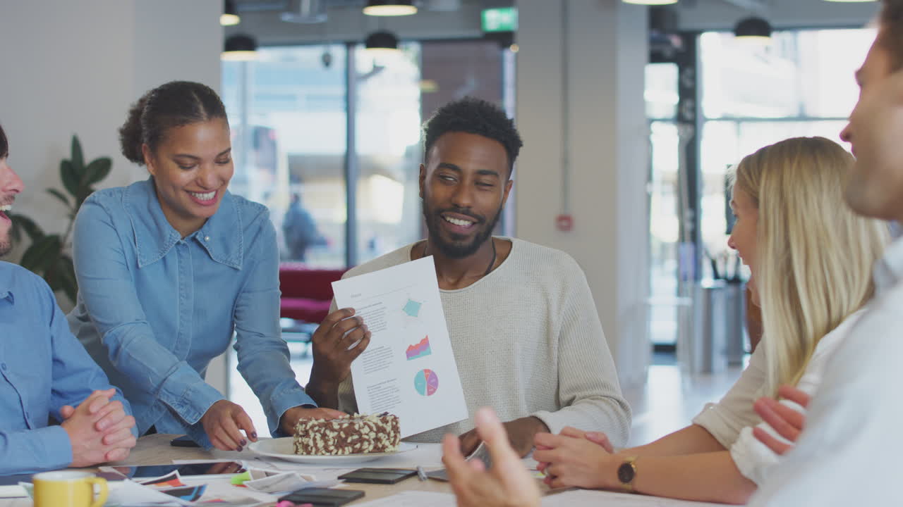 Colleagues Celebrating Businessman's Birthday At Meeting Around Table In Modern Open Plan Office