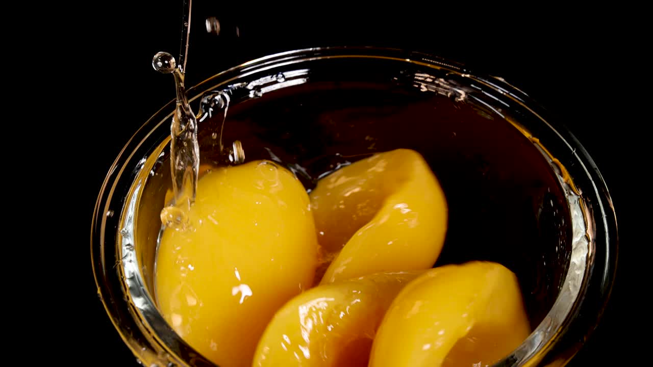 Halved yellow peaches are dropped into a clear glass bowl filled with syrup, captured in close-up with dramatic lighting against a black background