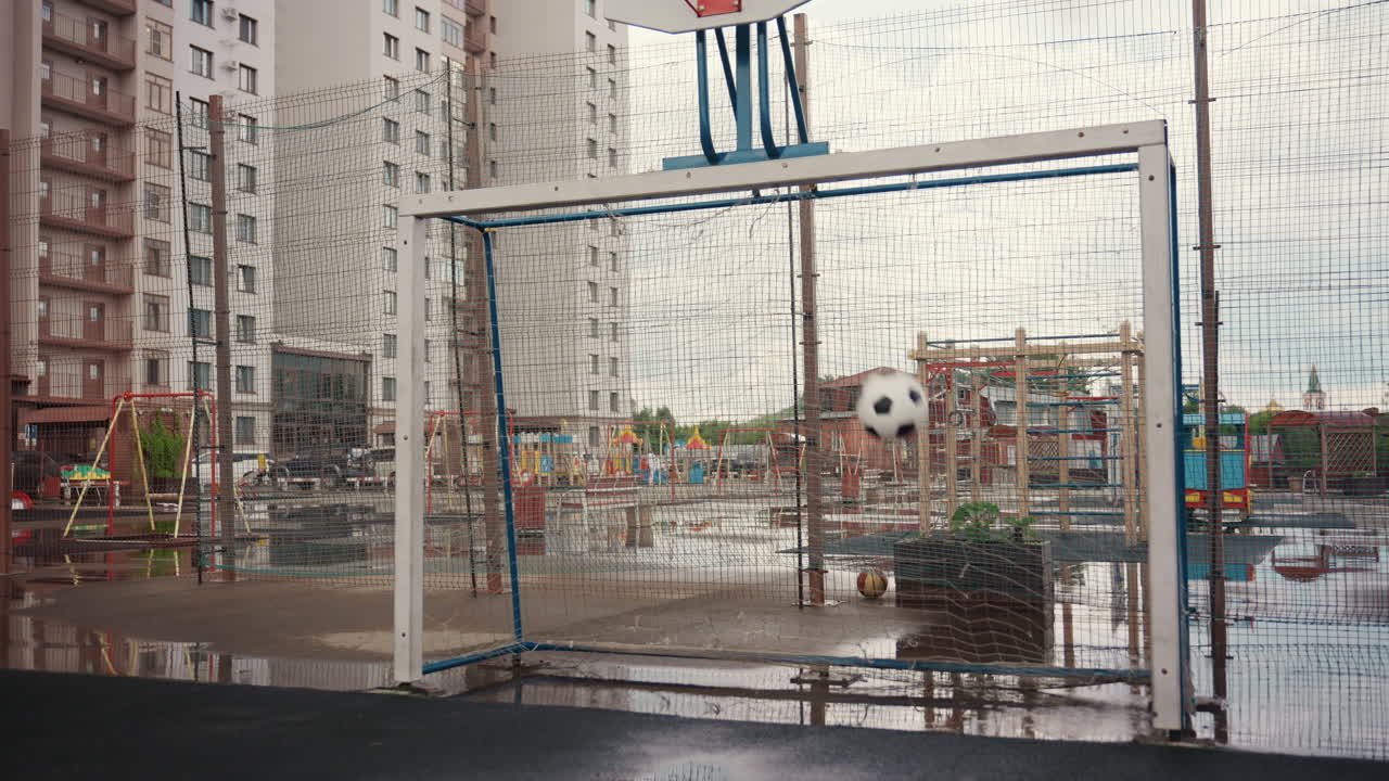 Regnvåt urban fotballmålscene, tom bane med vått nett, dammer på asfalt, høyhus i bakgrunnen, overskyet himmel som skaper en stemningsfull atmosfære, gjenværende fotball nær målet, i det fjerne