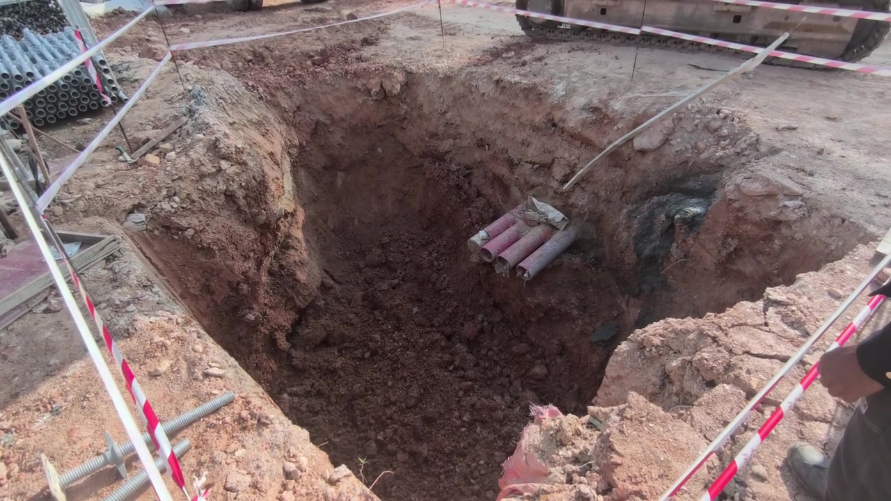 Bir ekskavatör, elektrik menholü kurulumu için derin bir çukur kazıyor. Kazara düşmeleri önlemek amacıyla parlak güvenlik bariyer bandı inşaat alanını çevreliyor.