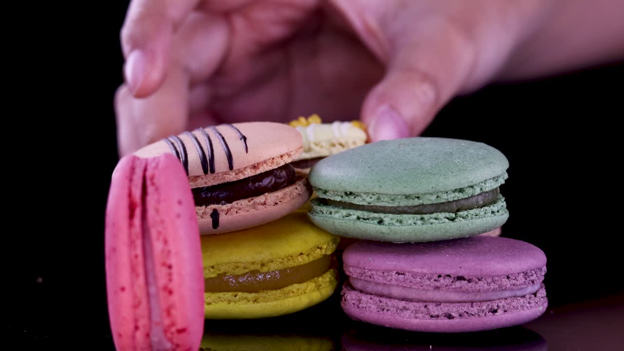 Hands carefully arrange assorted colorful macarons into a neat stack on a glossy black surface, under bright studio lighting with a close-up perspective