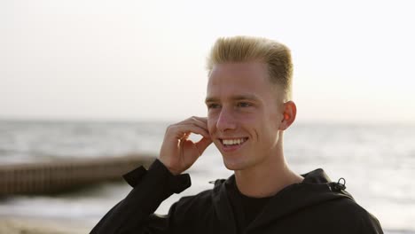 portrait of a young man who puts and earphone into his ear and listens to music against the backdrop of the waves of the sea at