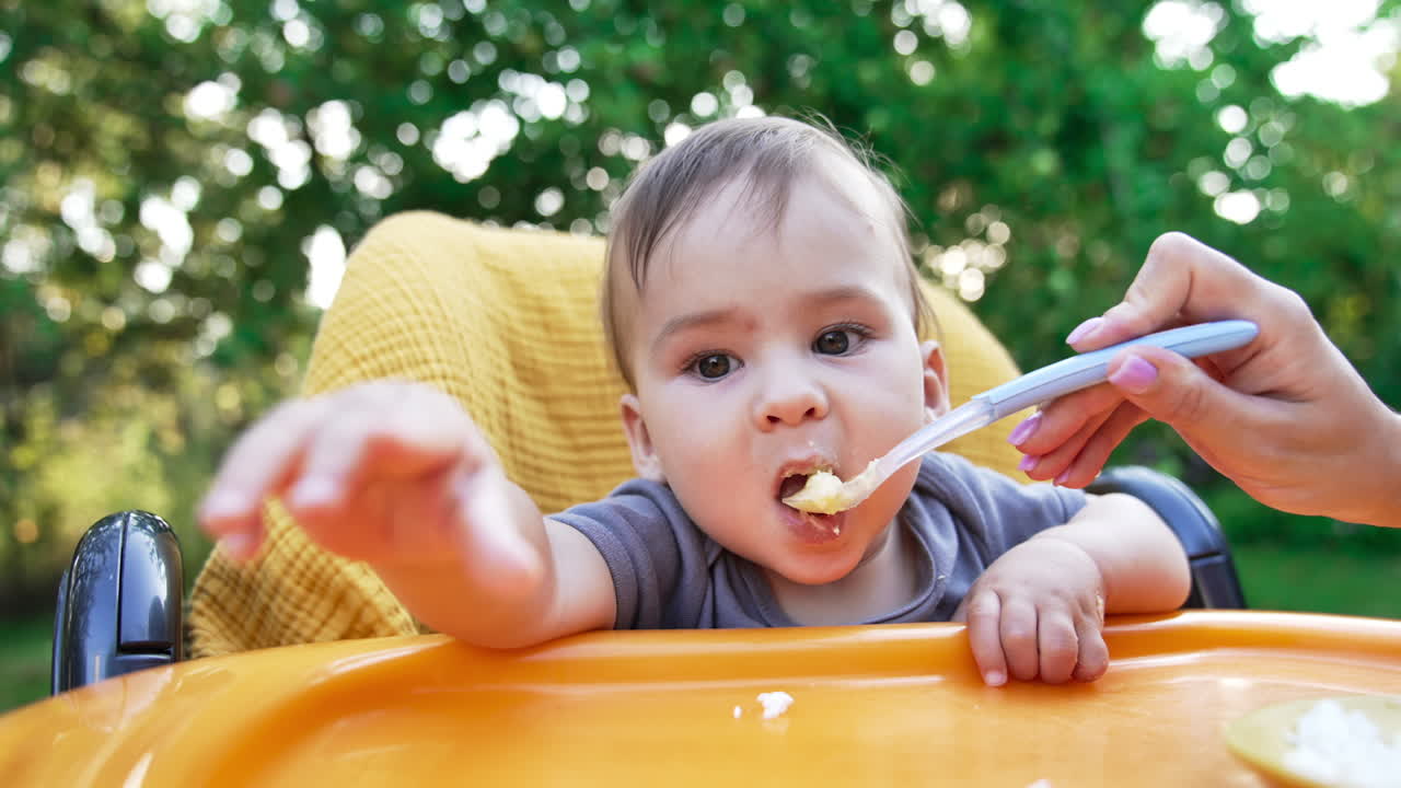 Feeding a fine curious baby outdoors. Little toddler sitting at yellow feeding table eats willingly from a spoon. Blurred nature at backdrop.
