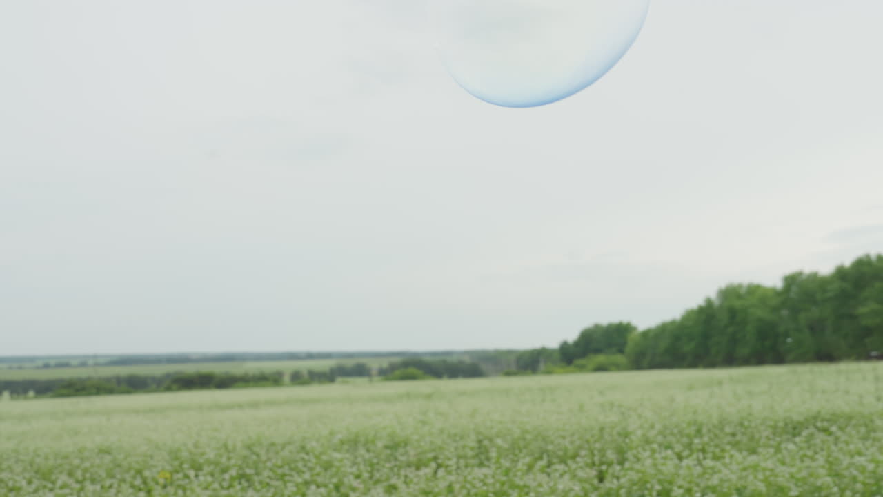 백인 여성이 붉은색 막대로 초원 위에서 거대한 비눗방울을 만들고, 부드러운 구름이 덮인 하늘 아래 넓게 펼쳐진 풍경과 멀리 보이는 나무들이 있는 가운데, 무지갯빛 구슬이 천천히 떠다니며 명상적인 야외 예술과 평온함을 연출합니다.