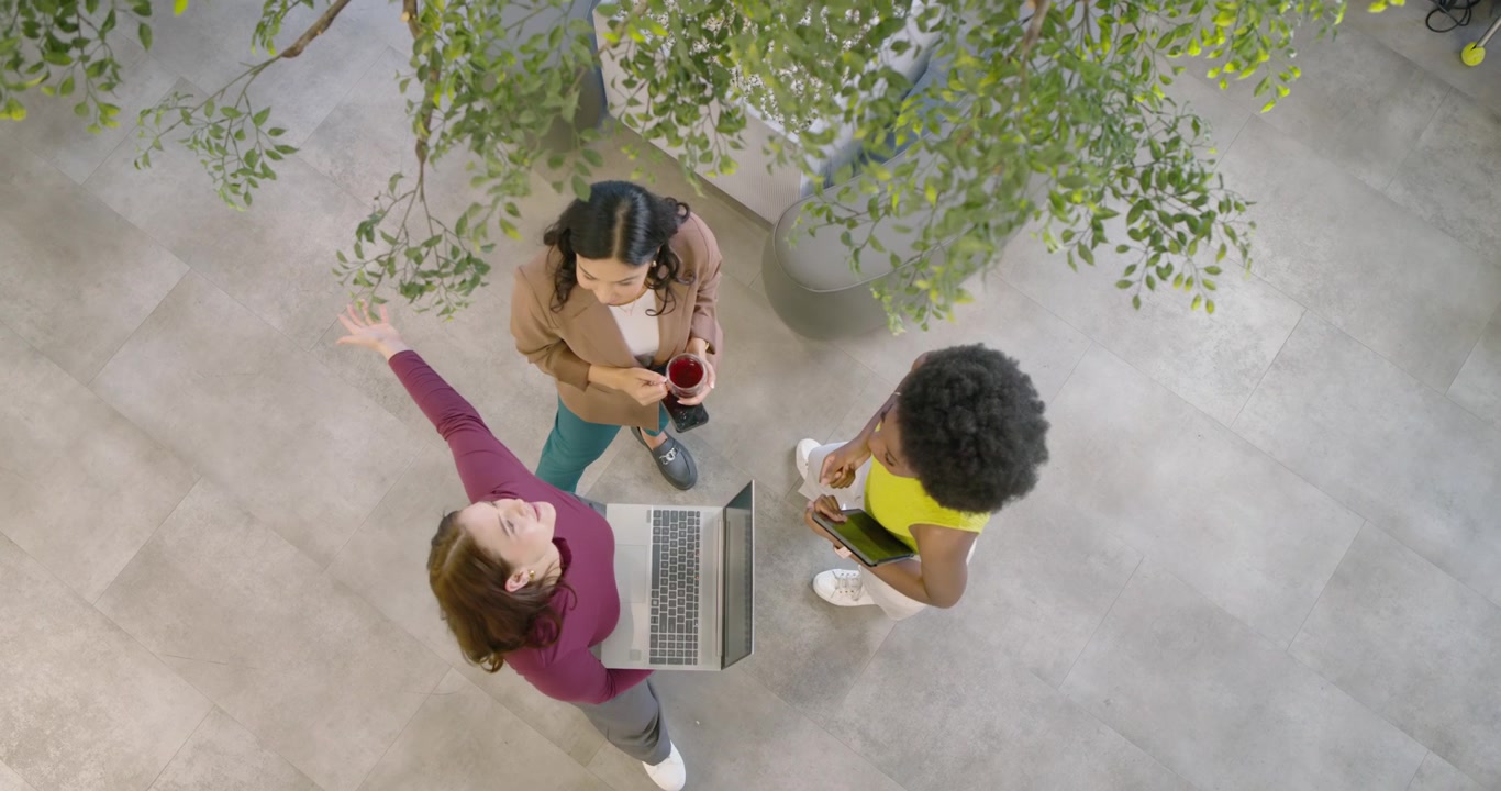 Diverse female coworkers gathering as presenter angling laptop in atrium, mug and tablet for review