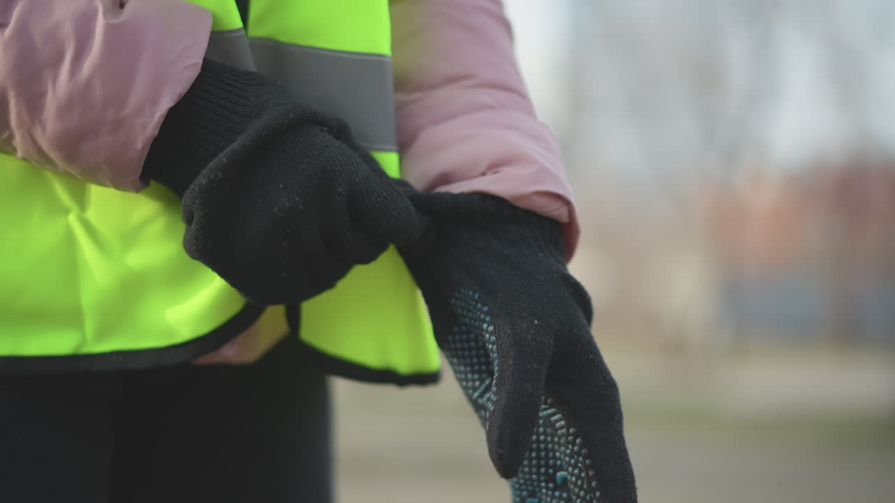 Close-up of person wearing bright neon yellow high-visibility safety vest with reflective stripes and pink padded jacket putting on black knitted gloves outdoors, preparing for work